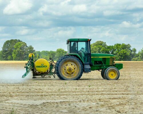 A green tractor pulls a yellow Redick sprayer attachment across a large partially tilled field under a cloudy sky The field is bordered by trees in the background