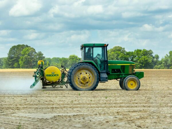 A green tractor pulls a yellow Redick sprayer attachment across a large partially tilled field under a cloudy sky The field is bordered by trees in the background